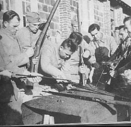 A group of men in military uniforms stand around a table outdoors, inspecting and cleaning rifles.