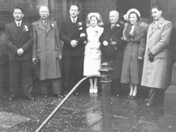 Six adults dressed formally stand in a line outdoors on a wet pavement, posing for a black-and-white group photograph.