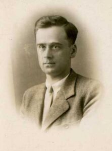 Black and white portrait of a young man wearing a suit and tie, looking slightly to the side with a neutral expression.