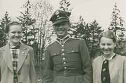 Three people pose outdoors; a man in a military uniform stands between two women dressed in coats, with trees in the background.