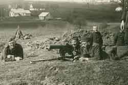 Three adults pose with a mounted machine gun in a rural field while a young child stands nearby; houses and trees are visible in the background.