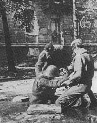 Three soldiers attend to a wounded comrade on the ground in an outdoor urban setting, with a building wall in the background.