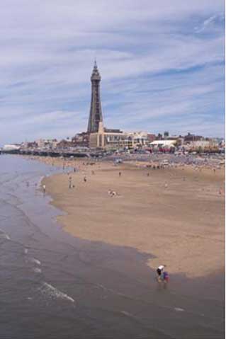 A sandy beach with scattered people, with a tall tower and buildings in the background under a partly cloudy sky.