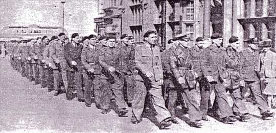 A group of uniformed soldiers march in formation down a city street, passing buildings, in a black-and-white historical photograph.