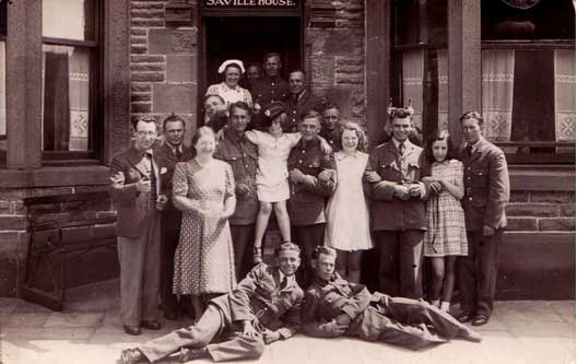 A group of men in military uniforms and civilians pose together outside a brick building labelled "Saville House.
