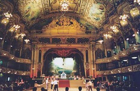 Ornate ballroom with elaborate ceiling frescoes, chandeliers, a large stage, and several people dancing on a polished floor.
