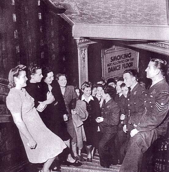 A group of men in military uniforms and women stand and talk in a crowded corridor; a sign behind them reads, "Smoking not allowed on the dance floor.