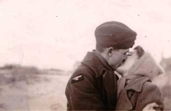 A person in a military uniform kisses a dog on the mouth outdoors.