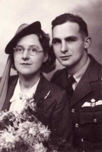 A black-and-white portrait of a woman in a hat and glasses holding flowers, seated next to a man in a military uniform with pilot wings.