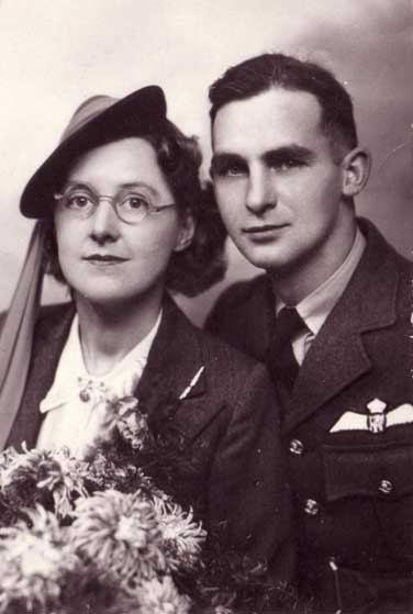A black-and-white portrait of a woman in a hat and glasses holding flowers, seated next to a man in a military uniform with pilot wings.