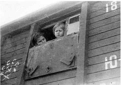 Two children look out from the window of a wooden train car. The exterior of the car has painted numbers and some writing.