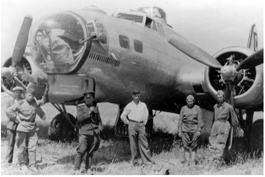 Five people stand in front of a vintage four-engine military aircraft on grass, with the plane's nose and propellers clearly visible.