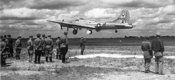 A group of soldiers watch as a B-17 bomber aircraft takes off or lands on a grassy airfield under a cloudy sky.