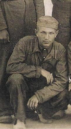 A young man in a military uniform crouches, looking at the camera. He wears a cap and is surrounded by other people, partially visible.