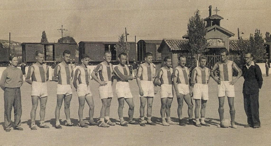A black-and-white photo of eleven men in striped football kits and two men in suits standing on a dirt pitch, with railway carriages and buildings in the background.
