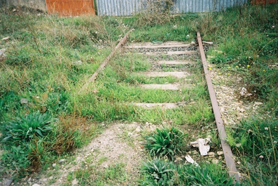 Abandoned railway tracks overgrown with grass and weeds, with some missing or damaged wooden sleepers, leading towards corrugated metal fencing.