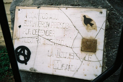 A weathered, cracked memorial plaque with faded Spanish text, a bird silhouette, and an anarchy symbol painted on the lower left-hand side.