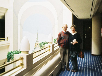 An elderly man and woman stand together in a corridor with blue carpet, white railings, and an arched window overlooking a building with a spire.