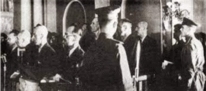 A group of men, some in military uniforms and some in suits, stand indoors during what appears to be a formal or official event.