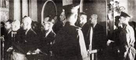 A group of men, some in military uniforms and some in suits, stand indoors during what appears to be a formal or official event.
