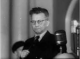 A man wearing glasses and a suit stands in front of a microphone, looking serious, in a black-and-white photo.
