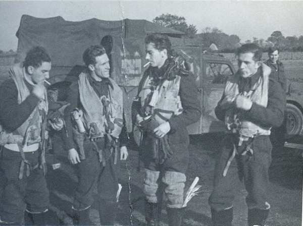 Four men in flight suits and life vests stand outdoors beside a military vehicle, engaged in conversation; one man smokes a cigarette.