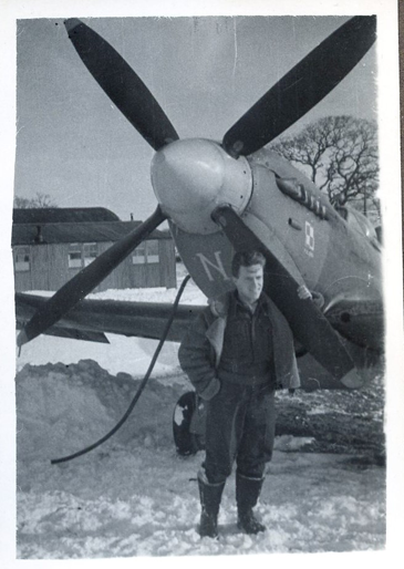 A man in a flight suit stands in front of a propeller aeroplane on snowy ground, with buildings and a tree visible in the background.