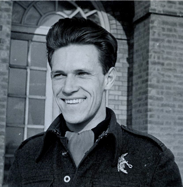 A man in a military-style jacket smiles outdoors in front of a brick building with a large arched window.