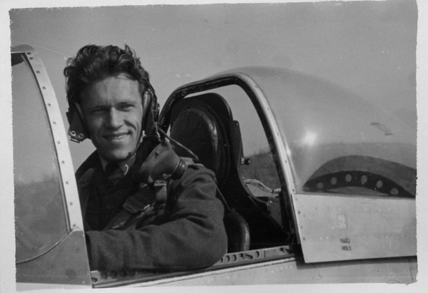 A man wearing a flight suit and headset sits in the cockpit of a military aeroplane with the canopy open, looking towards the camera and smiling.
