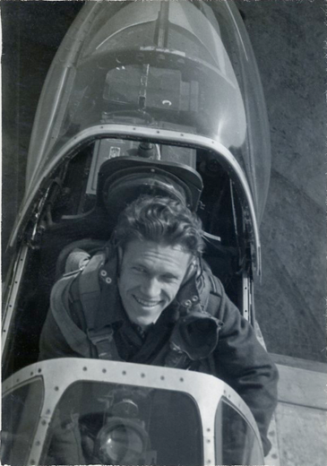 A man in a flight suit sits smiling inside the cockpit of a vintage aeroplane, viewed from above.