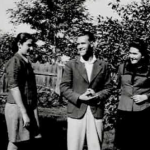 Three people stand outdoors in a garden or yard, smiling and talking, with trees and a wooden fence in the background.