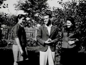 Three people stand outdoors in a garden or yard, smiling and talking, with trees and a wooden fence in the background.
