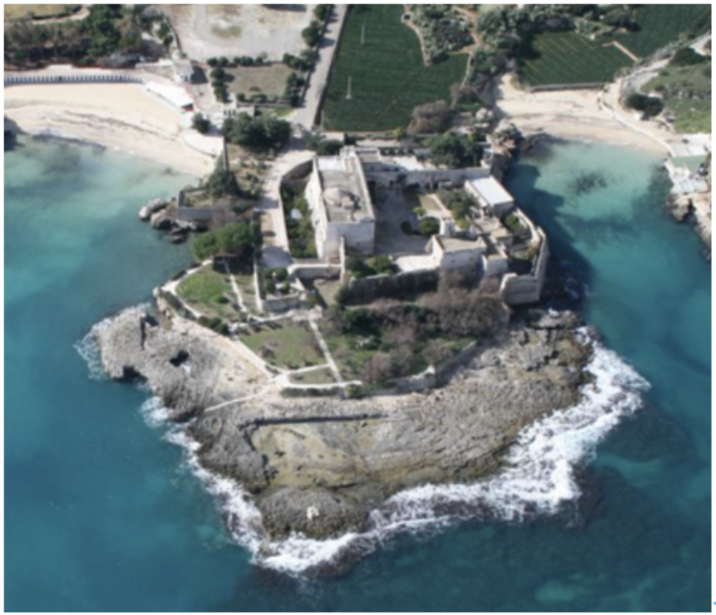 Aerial view of a stone fortress situated on a rocky island surrounded by turquoise sea, with beaches and greenery visible on the mainland in the background.