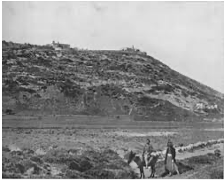 Two people with a donkey stand in a grassy area at the base of a rocky, sloping hillside with scattered buildings in the background.