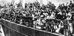 A crowded group of people stands and sits on the deck of a ship, many wearing head coverings and coats, appearing to be in transit or migration.
