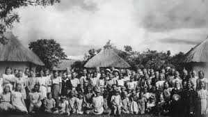 A large group of children pose outdoors in front of thatched-roofed huts, with trees and a cloudy sky in the background.