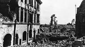 Black and white photo of bombed and ruined buildings with rubble covering the ground, illustrating heavy war damage in an urban area.