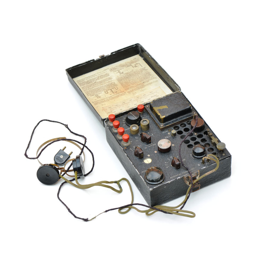 A vintage portable radio communication device with dials, buttons, wires, and headphones, open to show an instruction sheet inside the lid.