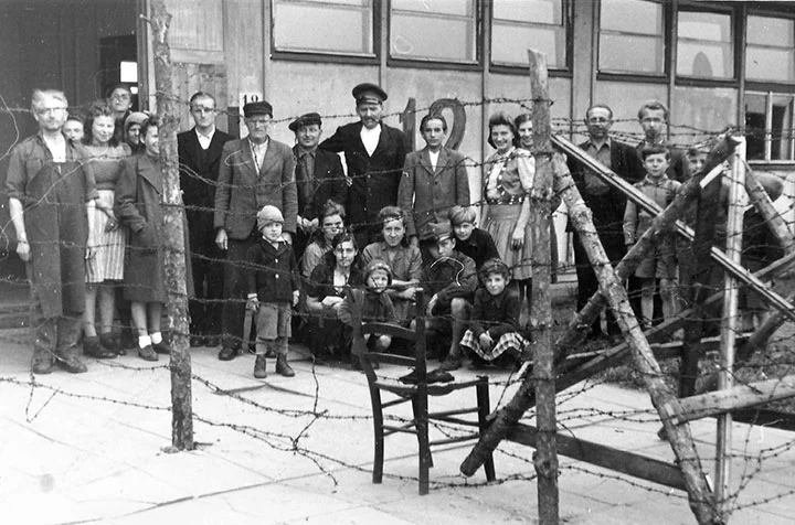 A group of men, women, and children stand and sit near a barbed wire fence and wooden barricade outside a building.