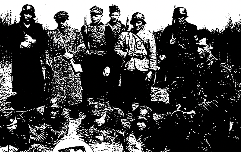 A group of soldiers in uniform, some standing and some lying on the ground with weapons, pose for a photo outdoors.