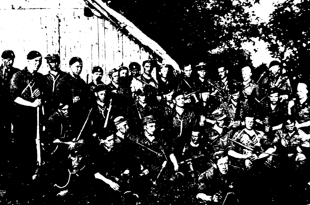A large group of armed men in uniform pose together outdoors in front of a wooden building and trees.