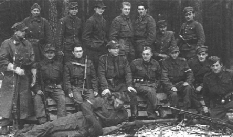 A group of uniformed soldiers pose for a black and white photo outdoors, with some seated on benches and others standing behind them; one person lies on the ground in front.