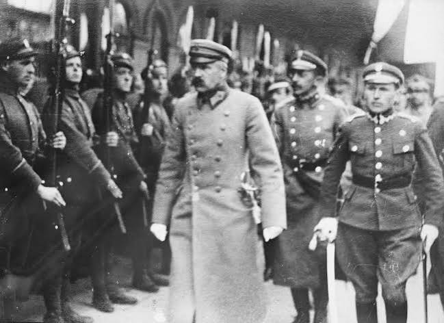 A group of military officers in uniform stand in formation, with one prominent officer walking in front, flanked by others.