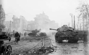 Two tanks and a damaged car sit on a debris-strewn street in a city with ruined buildings, whilst people walk and cycle nearby amid a hazy atmosphere.