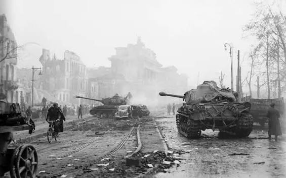 Two tanks and a damaged car sit on a debris-strewn street in a city with ruined buildings, whilst people walk and cycle nearby amid a hazy atmosphere.