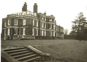 A large, historic, multi-storey brick mansion with many tall windows, chimneys, and a flat roof, surrounded by grass and trees, with steps leading up to the lawn.