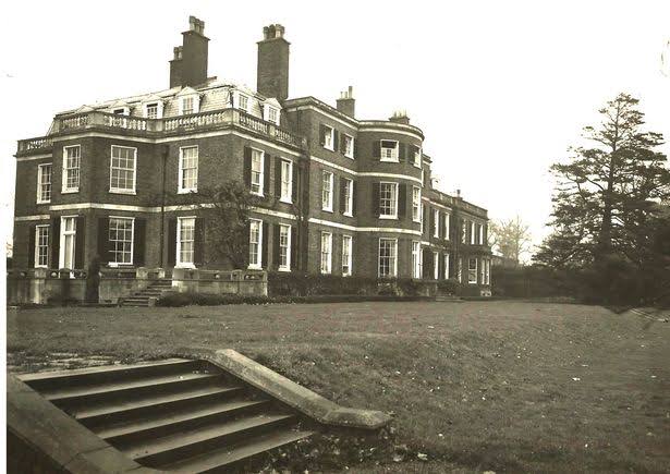Black-and-white photo of a large, three-storey Georgian-style mansion with tall chimneys, many windows, and a lawn with stone steps in the foreground.